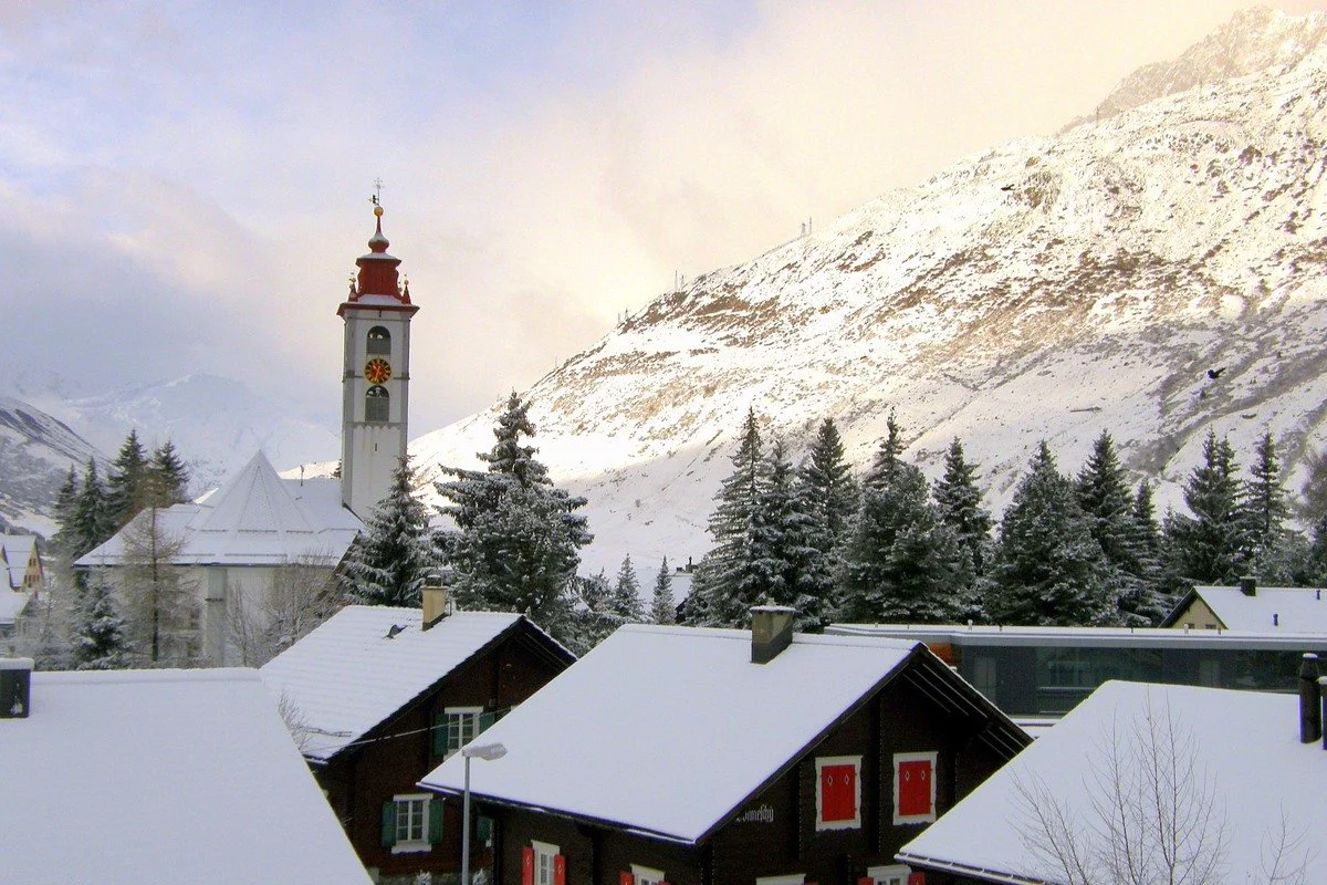 Snow-covered rooftops and evergreen trees in Andermatt mountain village with a church tower, snow-capped mountains in the background, and a cloudy sky.