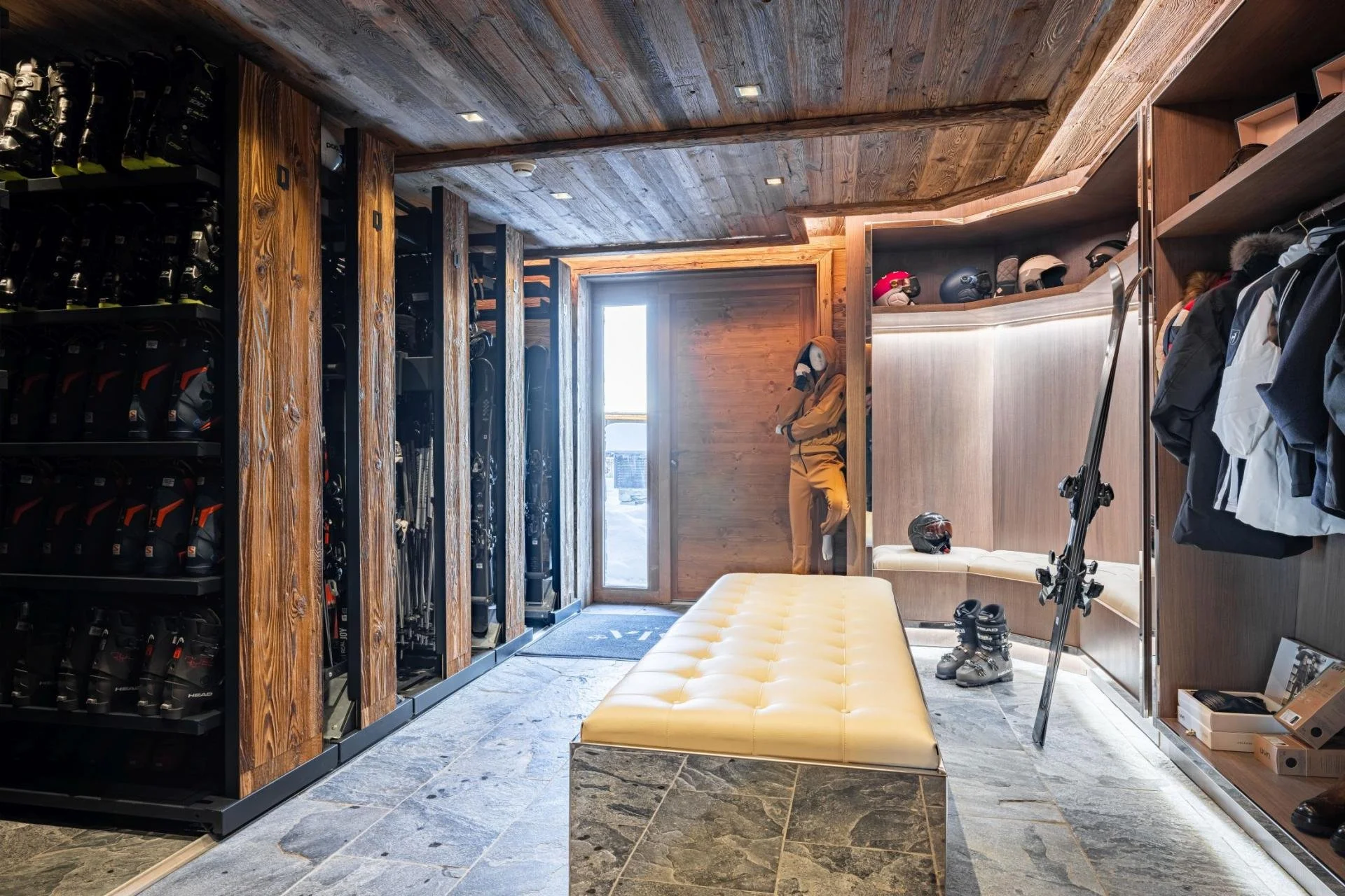 Ski gear and helmets on shelves, winter clothing on hooks, a scooter and ski boots on the floor, sunlight coming through a door, a person in ski gear in the background.