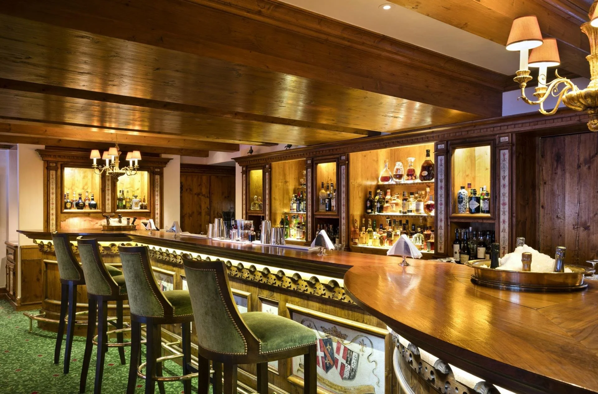 An elegant wooden bar with a polished countertop and a row of green upholstered barstools. Behind the bar, there are illuminated shelves displaying various bottles of liquor. The ceiling and walls are made of wood, and there are brass chandeliers wit