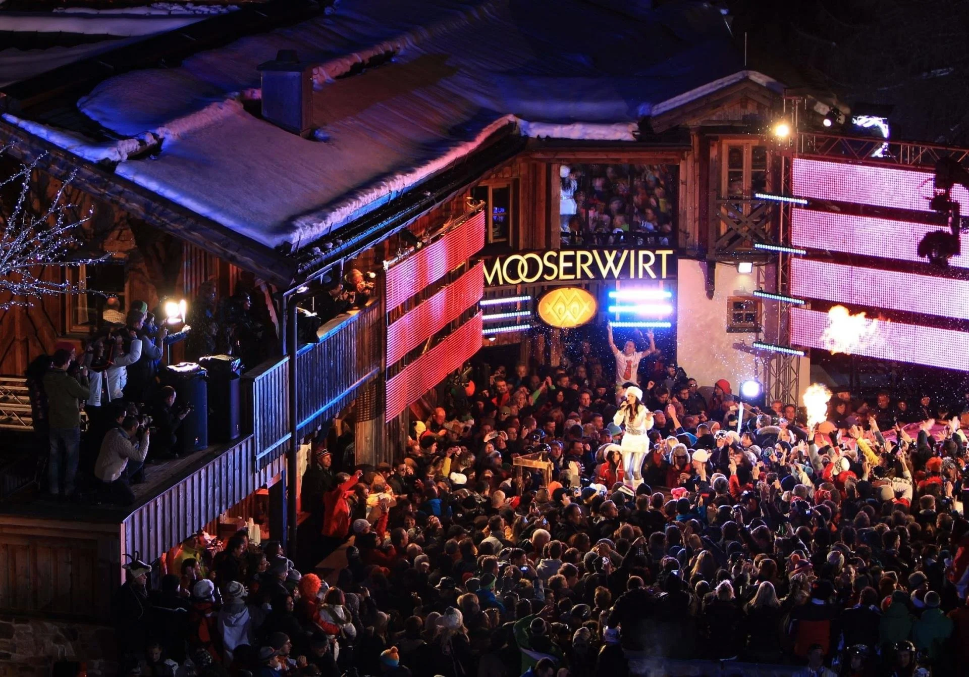 A crowded outdoor concert or event at night, featuring a stage with performers, bright lights, and a sign that reads 'MOOSEWIRT'. The audience is bundled in winter clothing, with some on a balcony and others on the ground, watching the performance.
