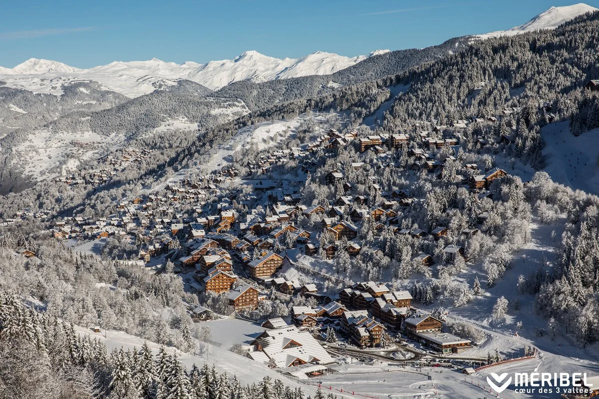 Snow-covered mountains and trees surround a village of Meribel with chalet-style houses on a hillside.