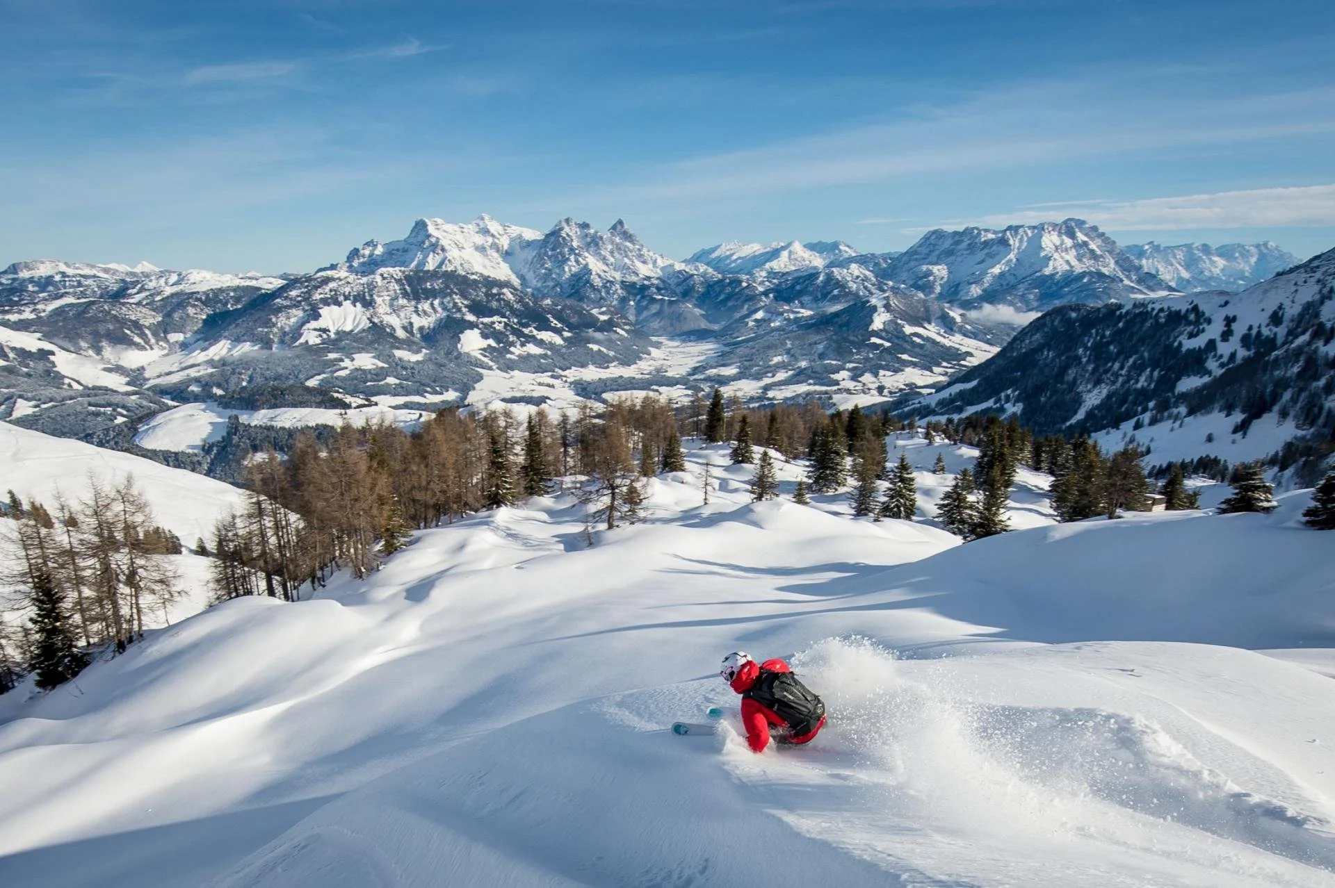 A person skiing down a snowy mountain slope in Kitzbuhel with snow-covered trees and mountains in the background on a clear sunny day.