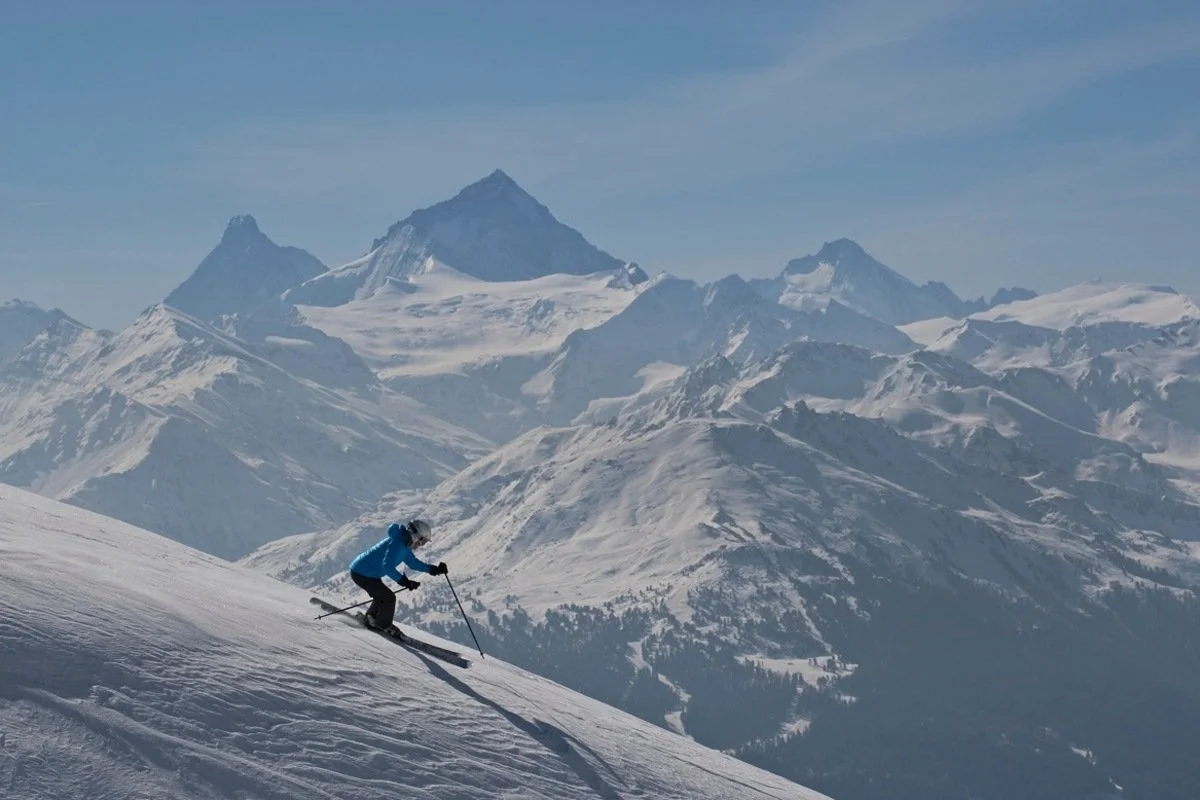A skier in a blue jacket and black pants skiing down a snowy mountain slope in Crans-Montana with snow-covered peaks and mountains in the background.
