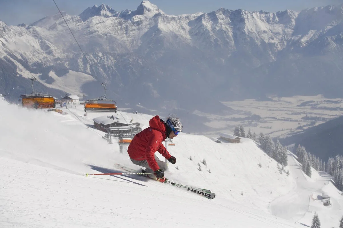 A skier in a red jacket and patterned helmet skiing down a snowy mountain slope with snow-covered mountains and ski lifts in the background.