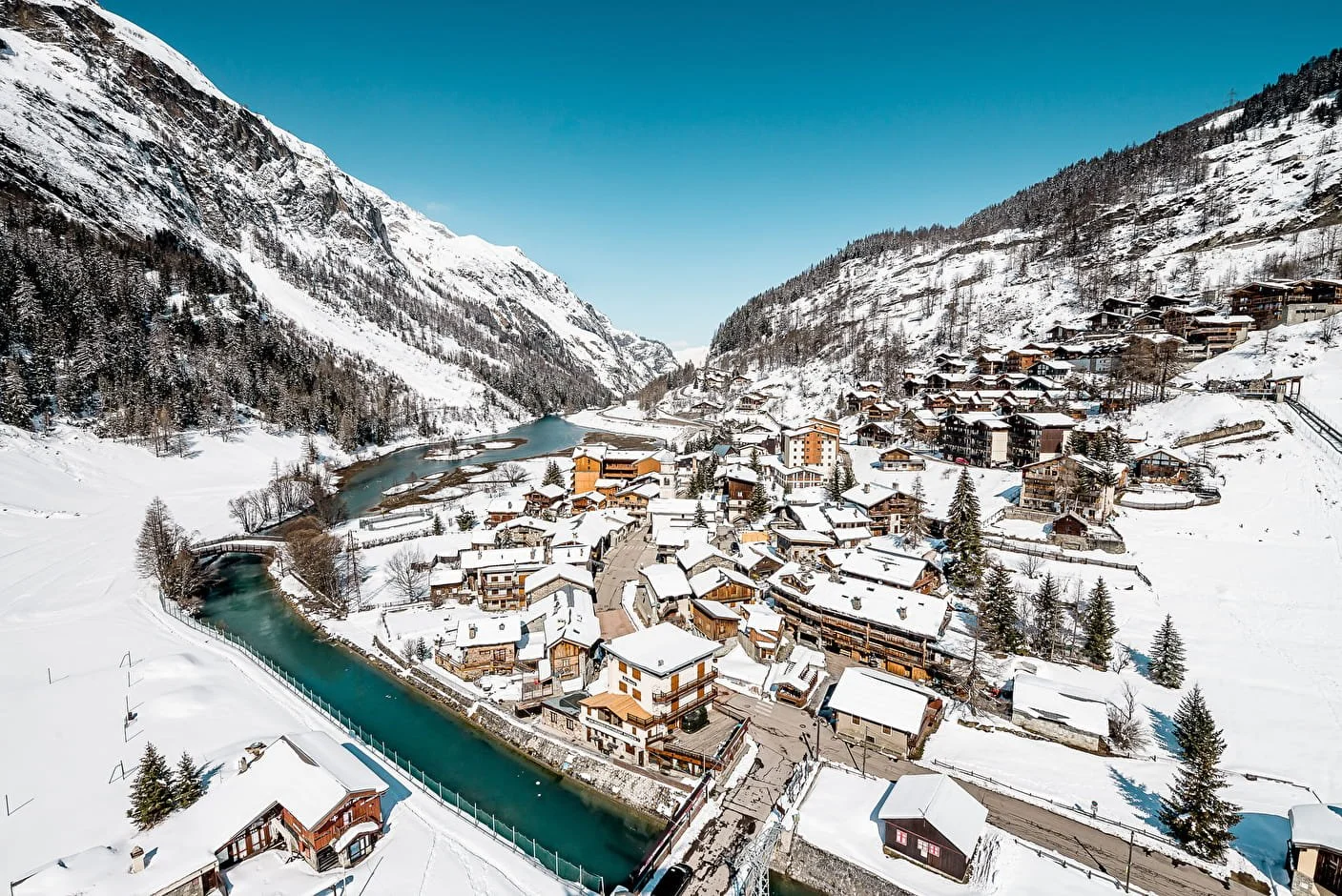 A snow-covered Tignes mountain village with a winding river, surrounded by tall, snow-capped mountains and pine trees.