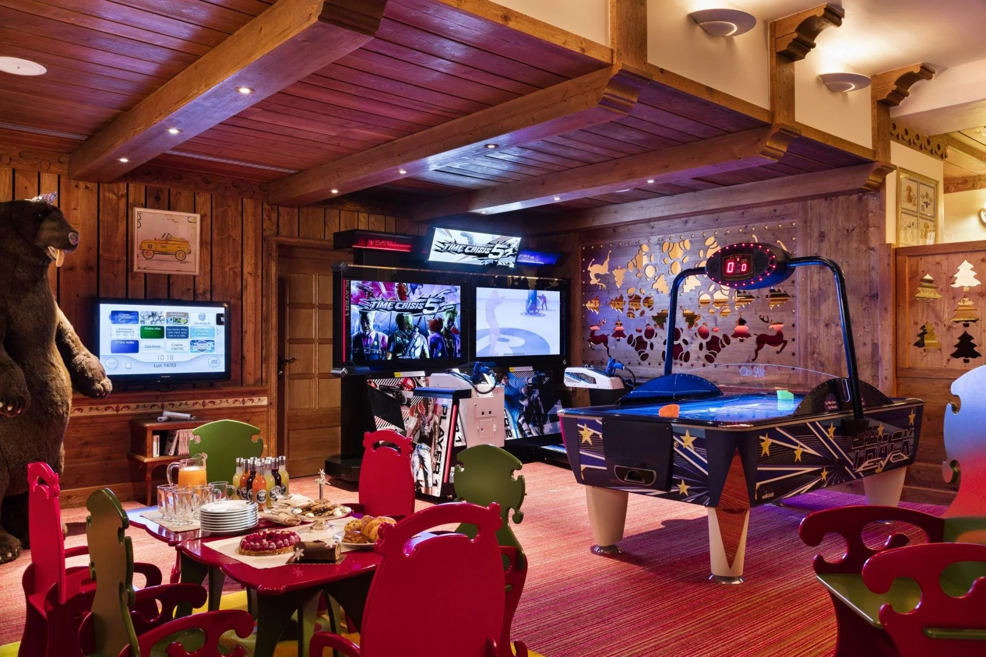 An indoor game room with a wooden ceiling and walls, featuring arcade machines, a digital scoreboard, and an air hockey table. In the foreground, there is a dining table set with plates and drinks, surrounded by colorful chairs.