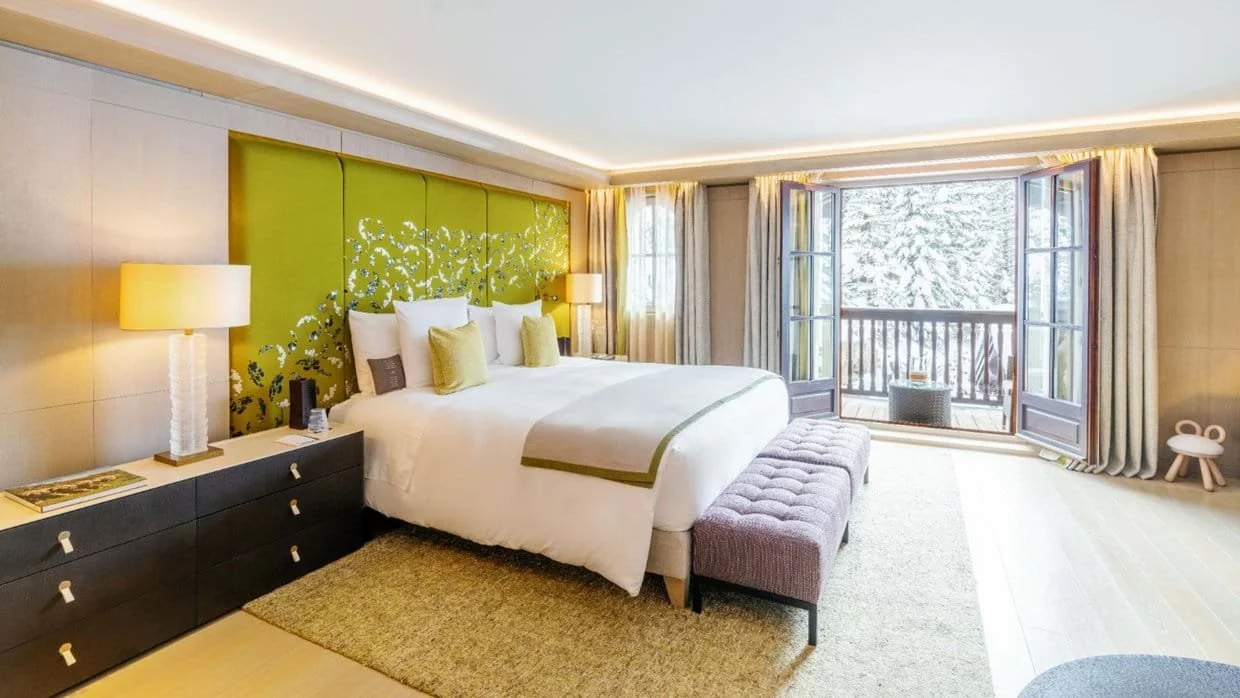 Hotel room with a large bed, green headboard, bench at the foot, bedside lamps, and a balcony door showing a snowy outdoor scene.