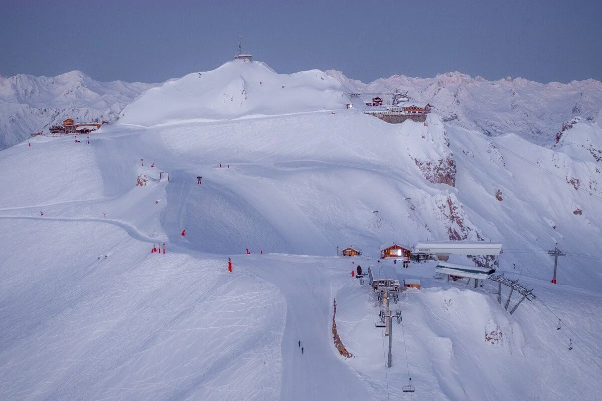 Snow-covered Courchevel 1850 mountain with ski lift, ski trail, and buildings at the top and bottom, during twilight.