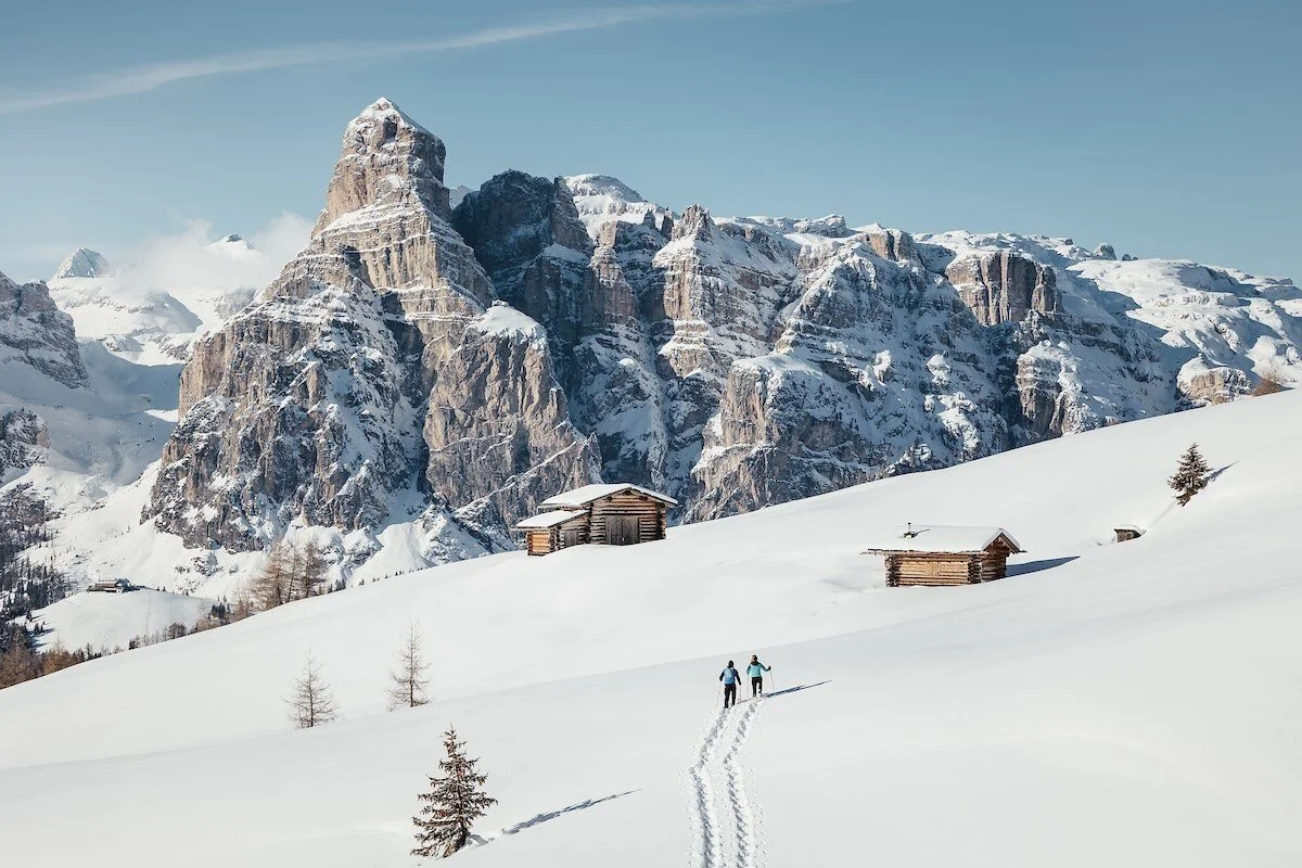 Two people cross-country skiing in a snowy landscape with small wooden cabins and a mountain range in the background.