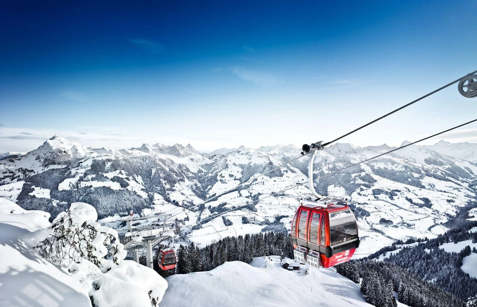 Cable cars flying over snow-covered Kitzbuhel mountains and trees in a winter landscape under a blue sky.