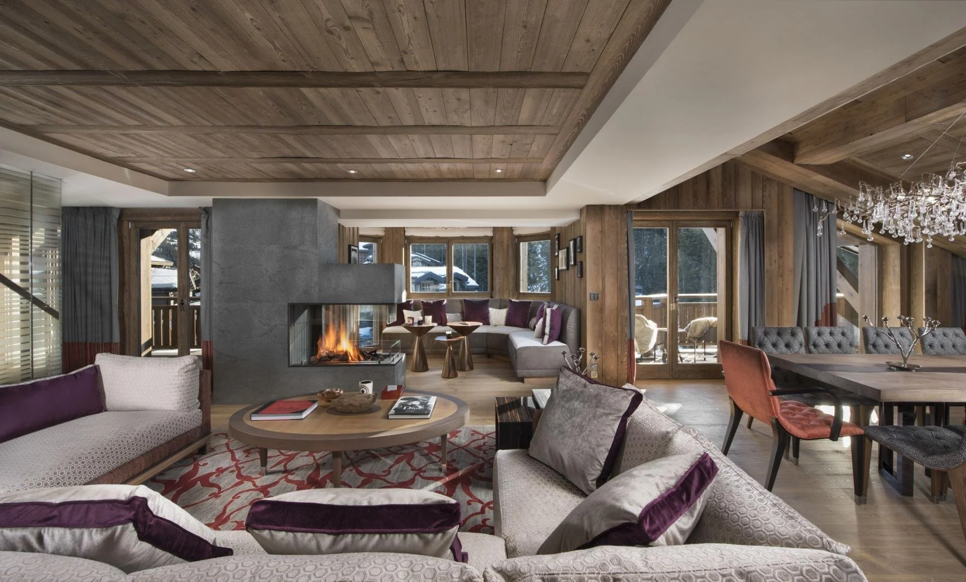 Cozy living room with wood-paneled ceiling and walls, a fireplace, large windows showing snowy outdoors, various sofas in neutral and purple tones, a dining table with a chandelier, and a balcony with snow outside.