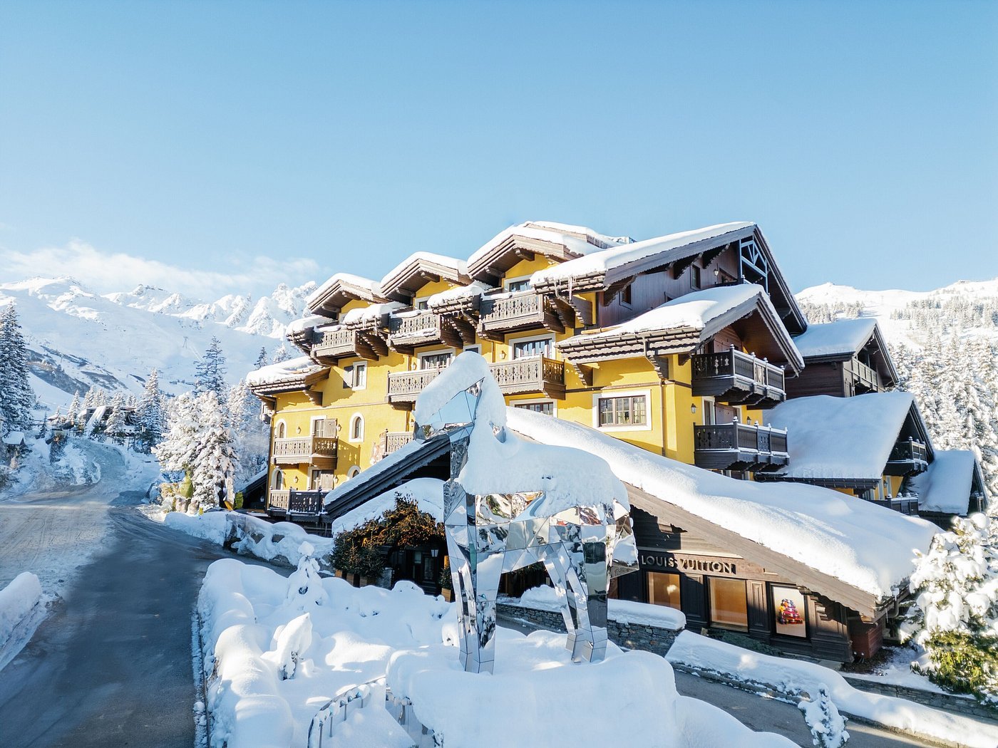 Yellow mountain lodge with snow-covered roofs in a snowy mountain landscape, with a reflective deer sculpture in the foreground.