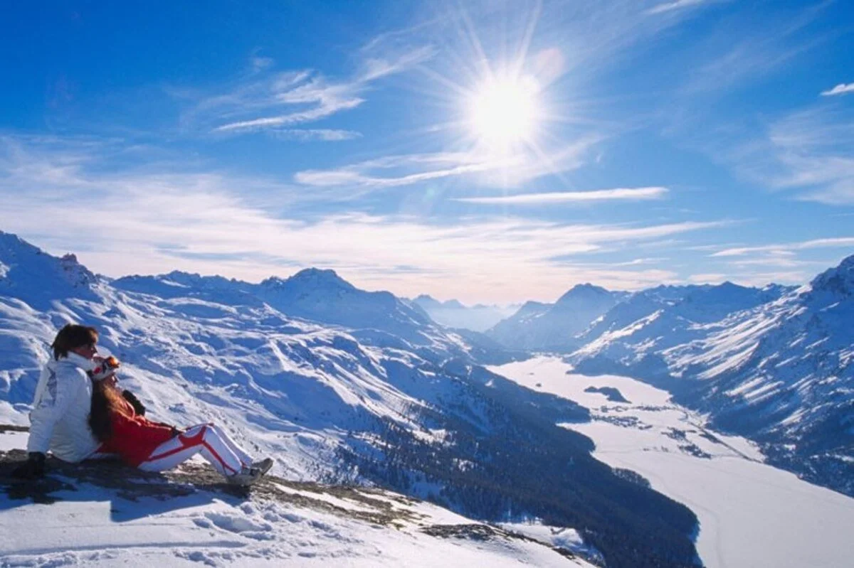 Two people sitting on snow-covered ground in St Moritz, overlooking a snowy mountain valley under a bright sun with a mostly blue sky and some clouds.