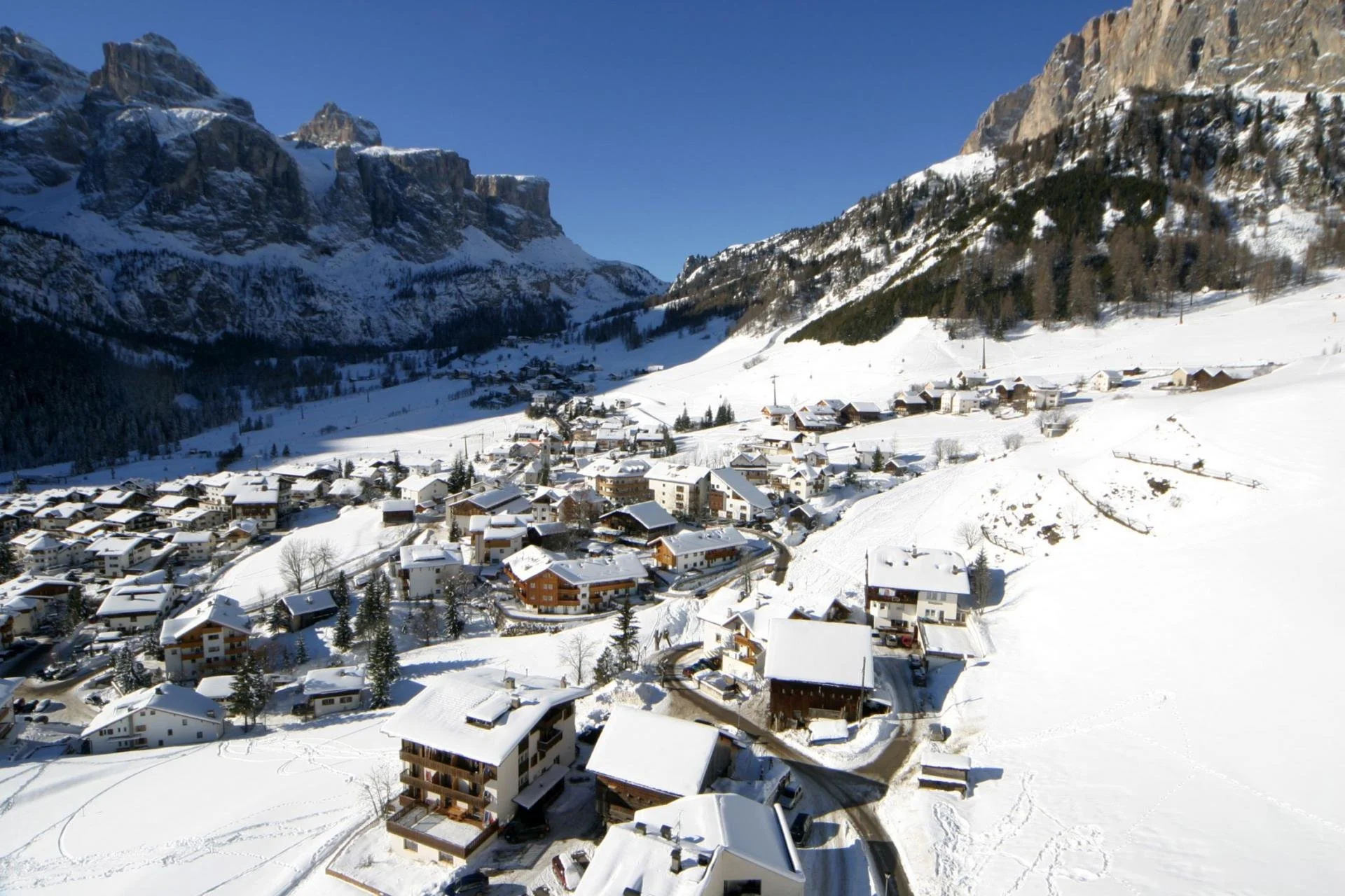 Snow-covered mountain village with houses and buildings, surrounded by mountains under a clear blue sky.