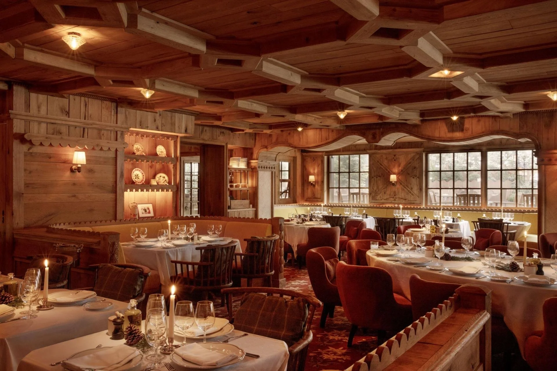 Interior of a wooden restaurant with decorated tables set for dining, large windows, and warm lighting.