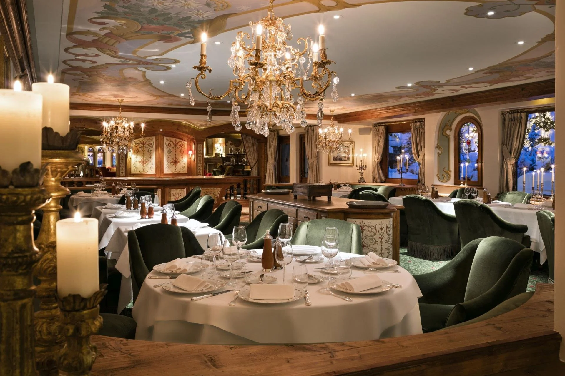 A luxurious dining room decorated in classic style with chandeliers, candles, and elegant table settings for a formal meal, with windows showing a snowy outdoor scene.