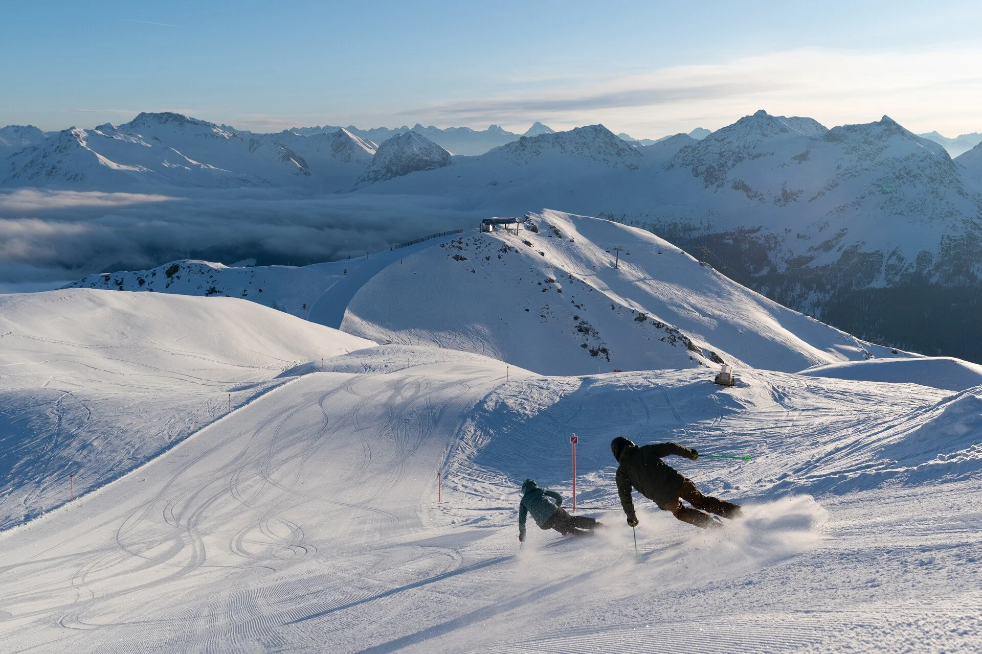 Two skiers descending a snowy mountain slope in Arosa with snow-covered mountains and a cloudy sky in the background.