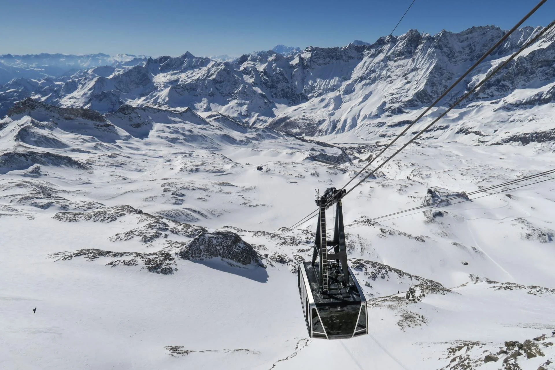 A ski lift gondola descending over snow-covered mountain landscape with rugged peaks in the background.