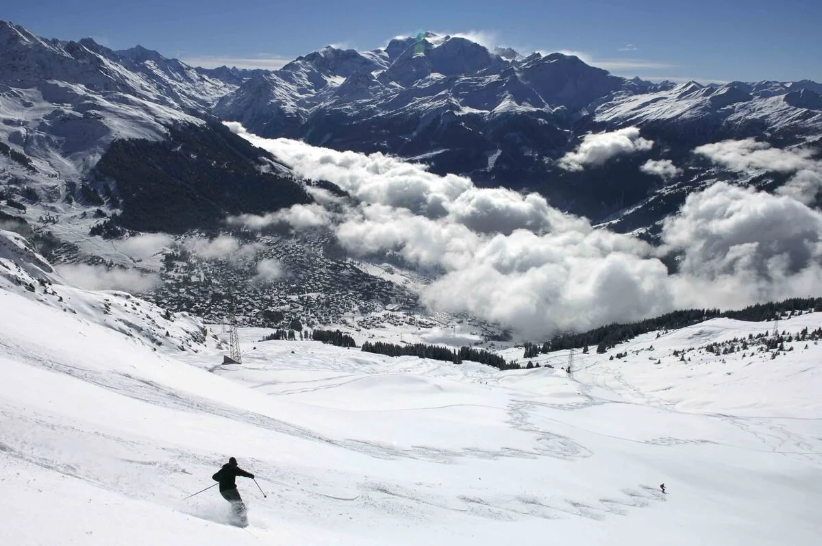 A skier in black gear skiing on snow-covered mountains in Verbier with a backdrop of tall, snowy peaks, cloudy sky, and a distant view of a valley and town below.