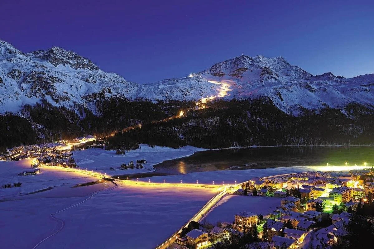 Snow-covered St Moritz mountain landscape at dusk with a small town at the foot, illuminated by streetlights and building lights, and a winding road through the snow.