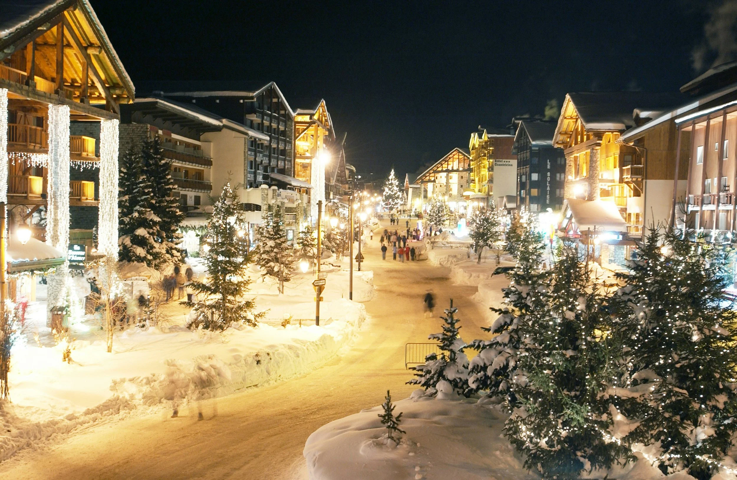 Night view of a snowy Christmas market street in Val d'Isere with decorated trees, illuminated buildings, and people walking.