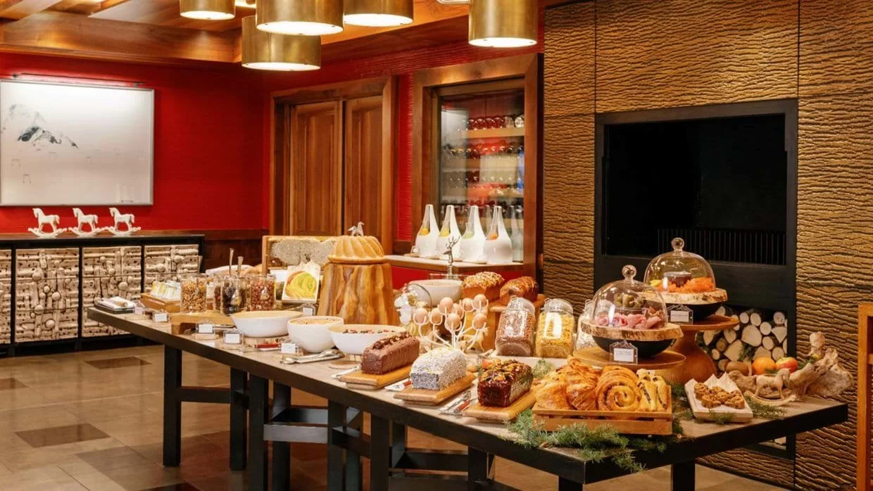 A dessert and food display table in a cozy room with wood interior, featuring cakes, pastries, jars of toppings, and decorative bottles.