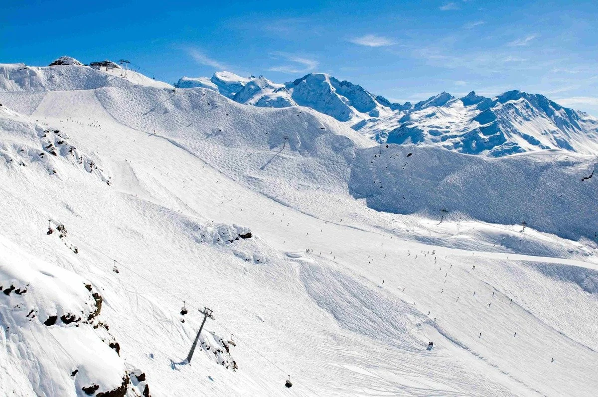 Snow-covered Verbier mountain ski resort with ski lifts and skiers on the slopes, mountain peaks in the background under a blue sky.