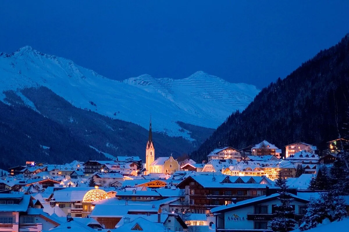 Snow-covered Ischgl alpine village in the mountains at dusk, with illuminated buildings and a prominent church steeple.