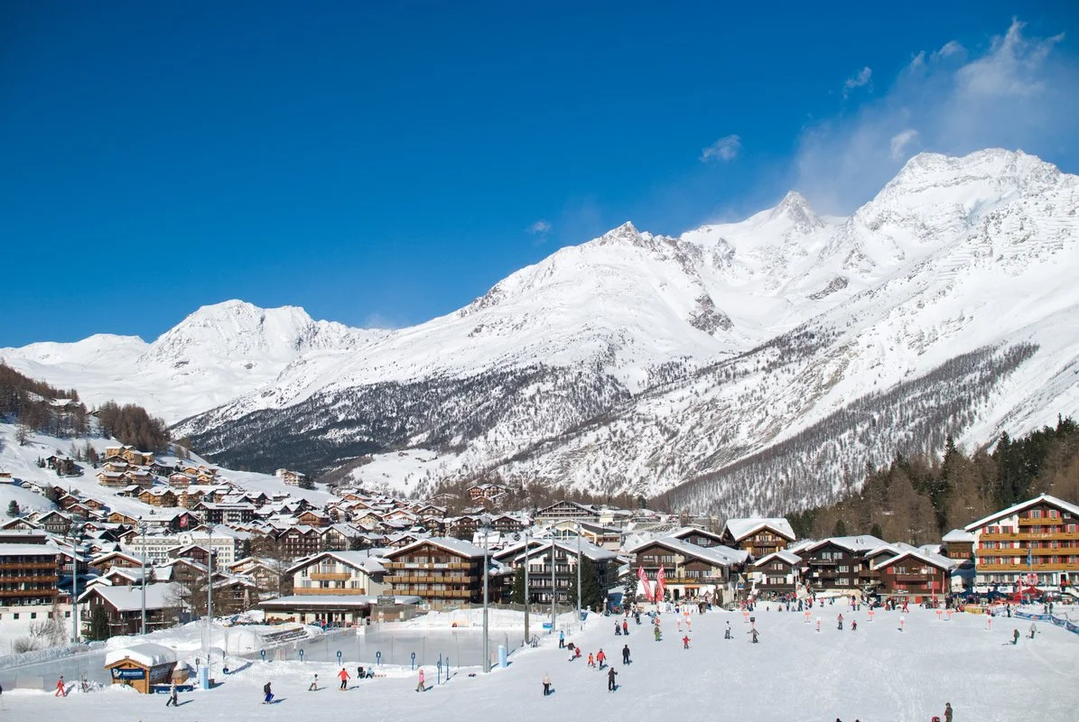 A snowy Saas-Fee mountain village with ski slopes and skiers, surrounded by tall snow-covered mountains under a clear blue sky.