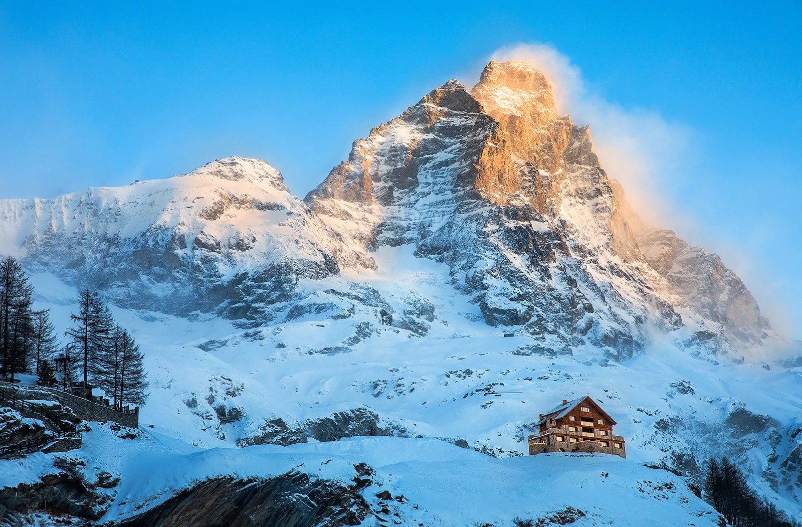 Snow-covered mountain with a house on a slope, trees, and an orange hue sunset in the background.