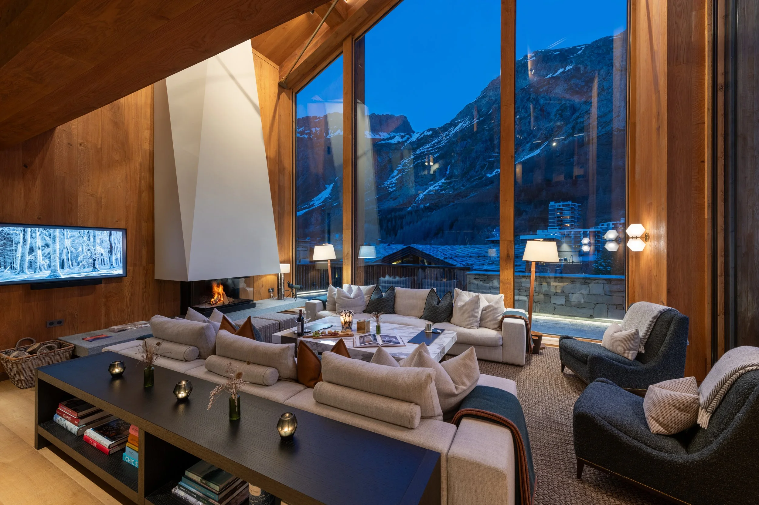 Living room with large floor-to-ceiling windows showing snowy mountains and a mountain town view. The room has a beige sectional sofa, modern chairs, a black coffee table with magazines, and a wood-paneled wall with a television and fireplace.
