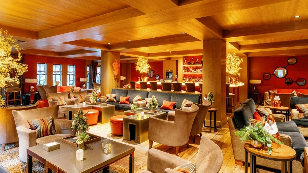 Interior of a cozy, warmly lit lounge with plush chairs, small tables, and holiday decorations including small Christmas trees and ornaments.