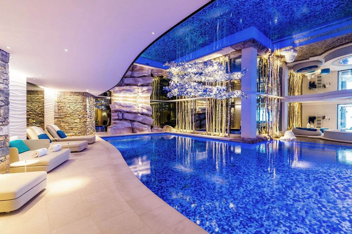 Indoor luxurious swimming pool area with modern decor, rock walls, decorative tree, and blue lighting.