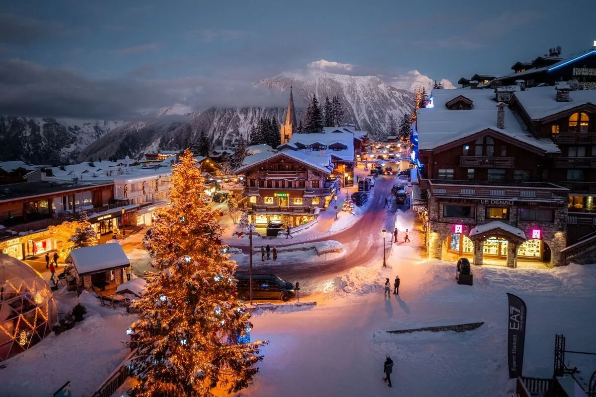 Snow-covered Courchevel 1850 square at dusk with illuminated Christmas tree, surrounding shops, and mountain backdrop.