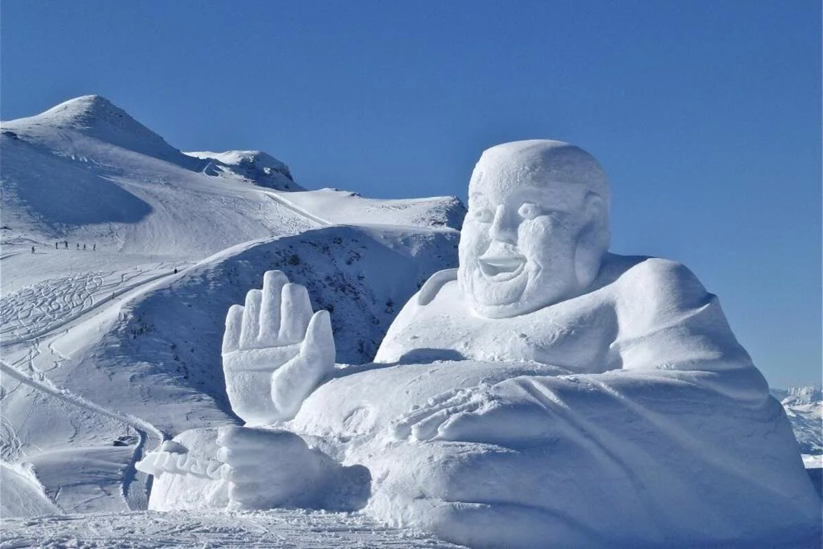 A massive snow sculpture in Arosa of a smiling man making a peace sign with his right hand, set against a snowy mountain landscape under a clear blue sky.