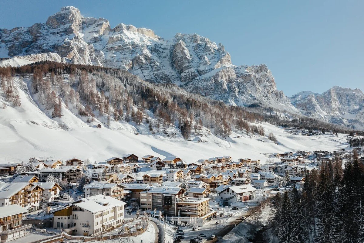 Snow-covered mountain village with wooden houses and trees set against a background of tall, rugged, snow-capped mountains and a clear blue sky.