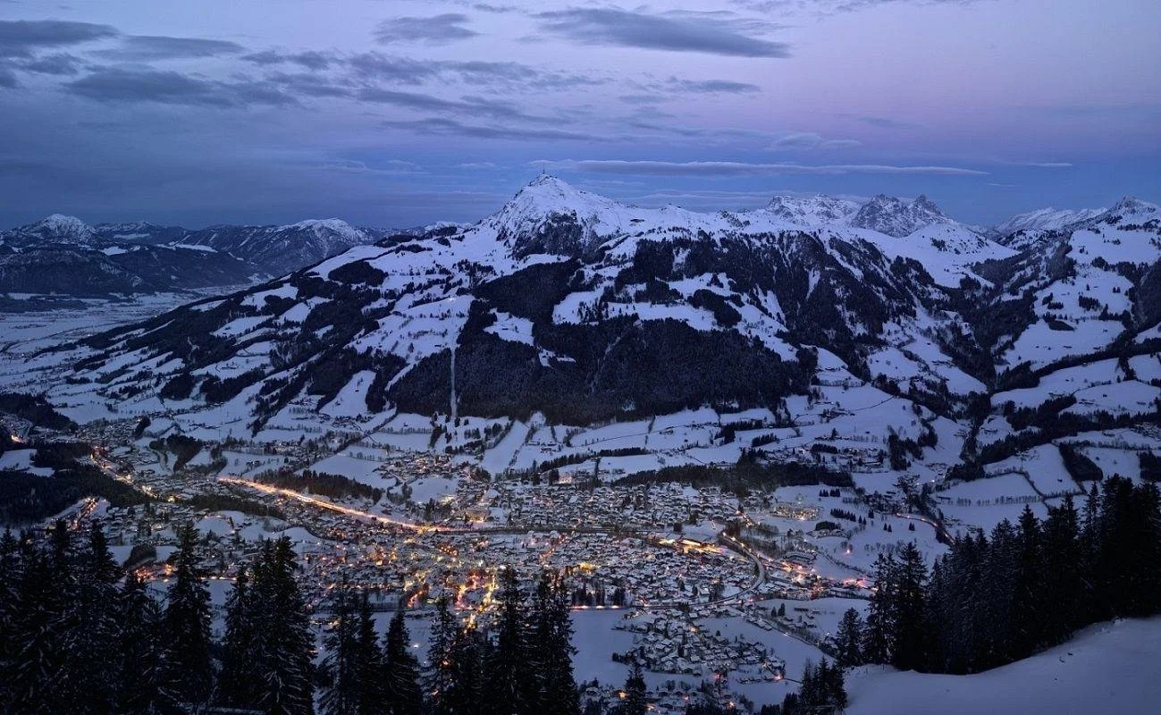 A snow-covered Kitzbuhel mountain village at dusk, with a tall mountain peak in the background, and the village lights illuminating the snow and trees.