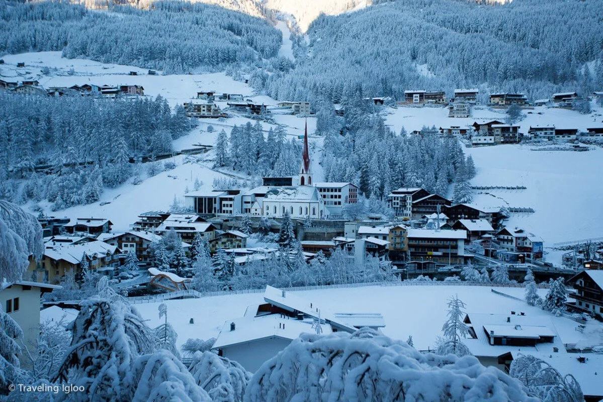Snow-covered Solden village with houses, trees, and a church with a tall steeple in a mountainous area.