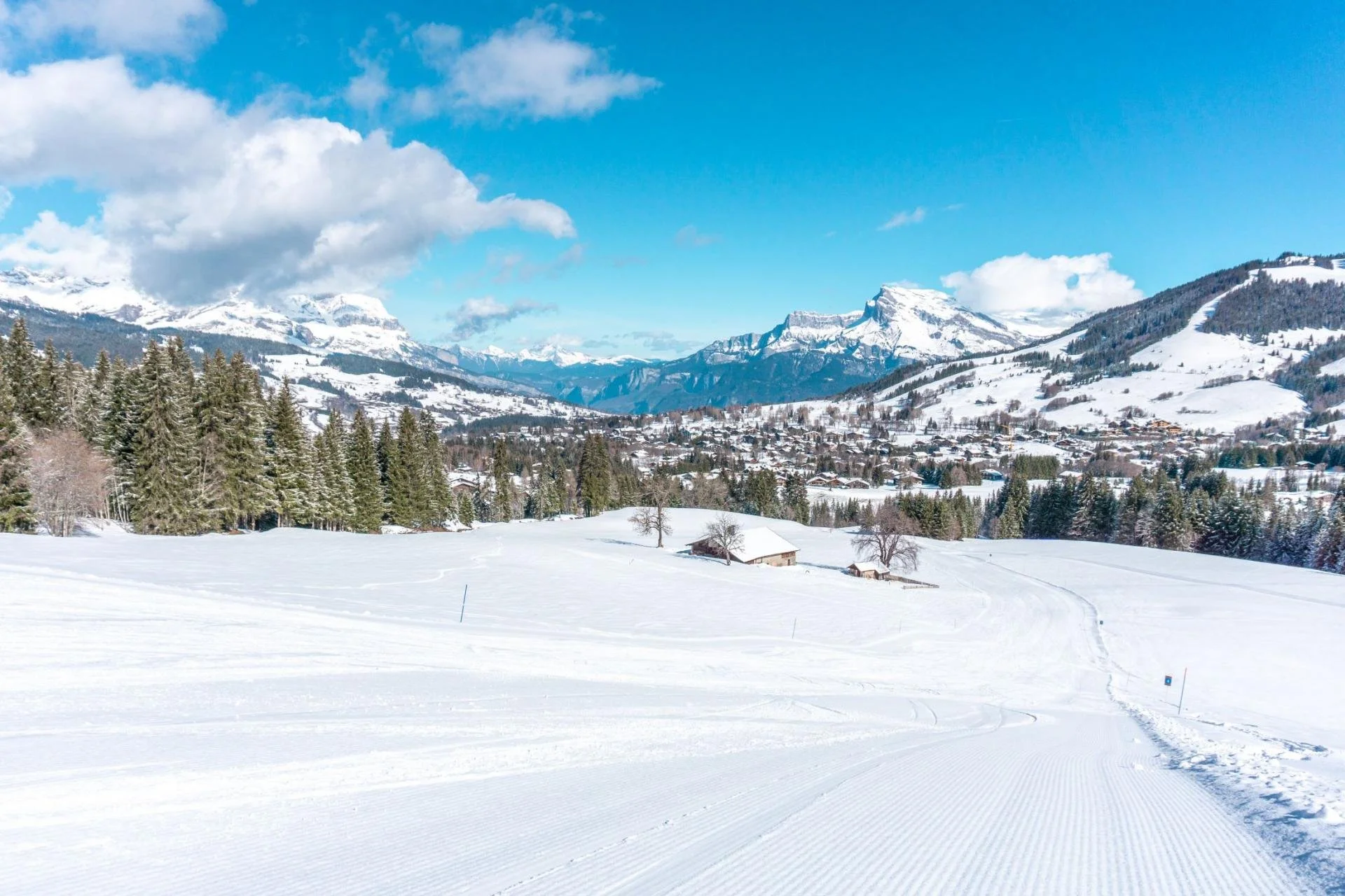 Snow-covered Megeve landscape with mountains in the background, a small village, and ski trails in the foreground on a clear, sunny day.