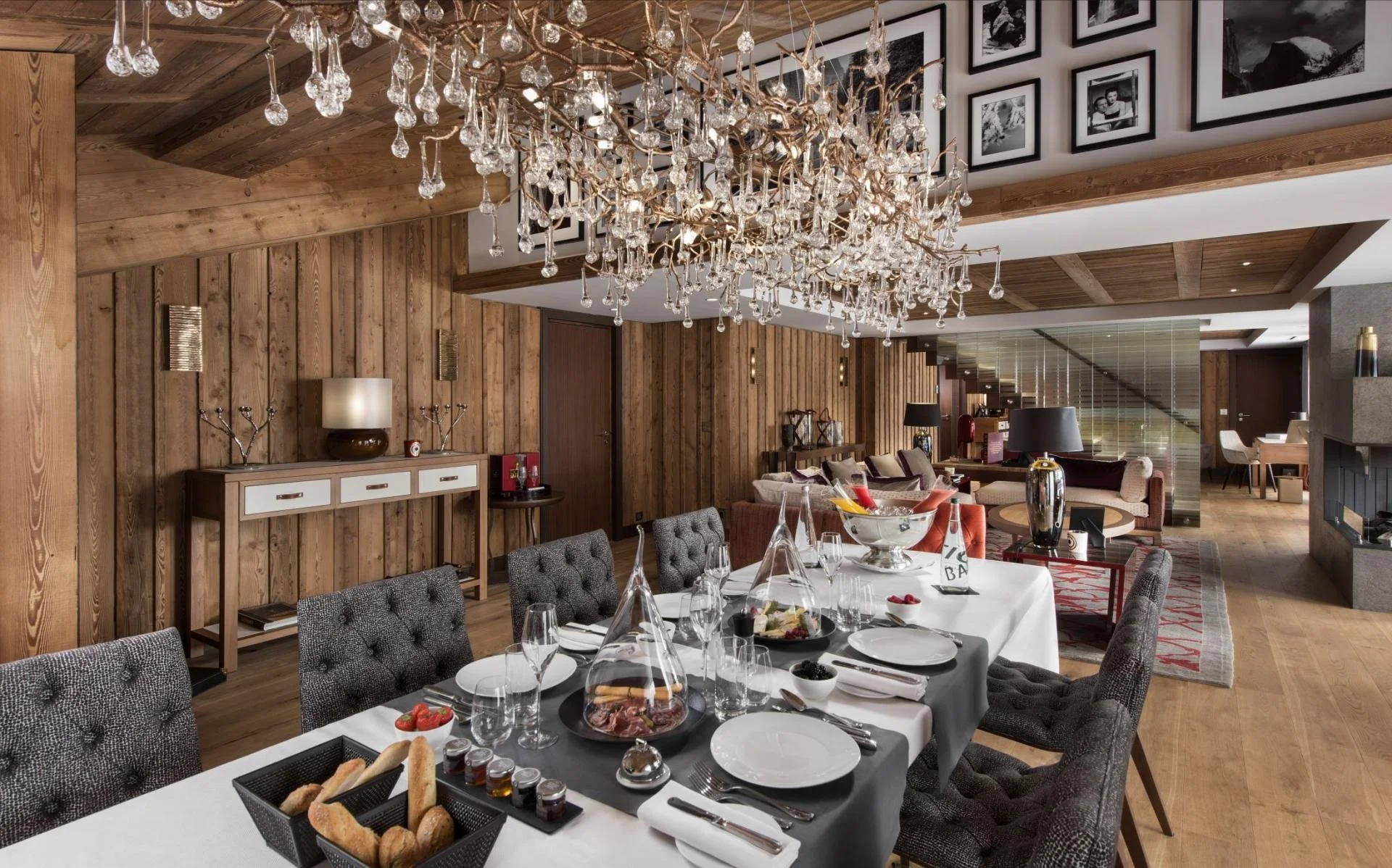 A cozy dining room with a large, elegant table set with wine glasses, plates, and a bread basket. The room features wooden wall paneling, a modern chandelier decorated with crystal drops, and framed black-and-white photographs on the wall. In the bac