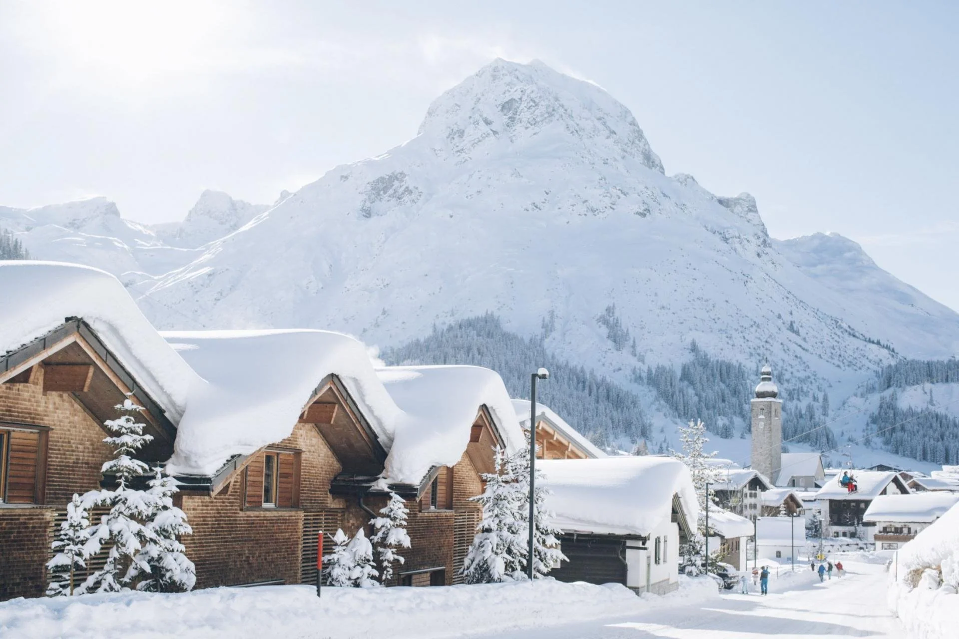 Snow-covered chalet-style buildings in Lech with a mountain in the background and people walking in the snow.