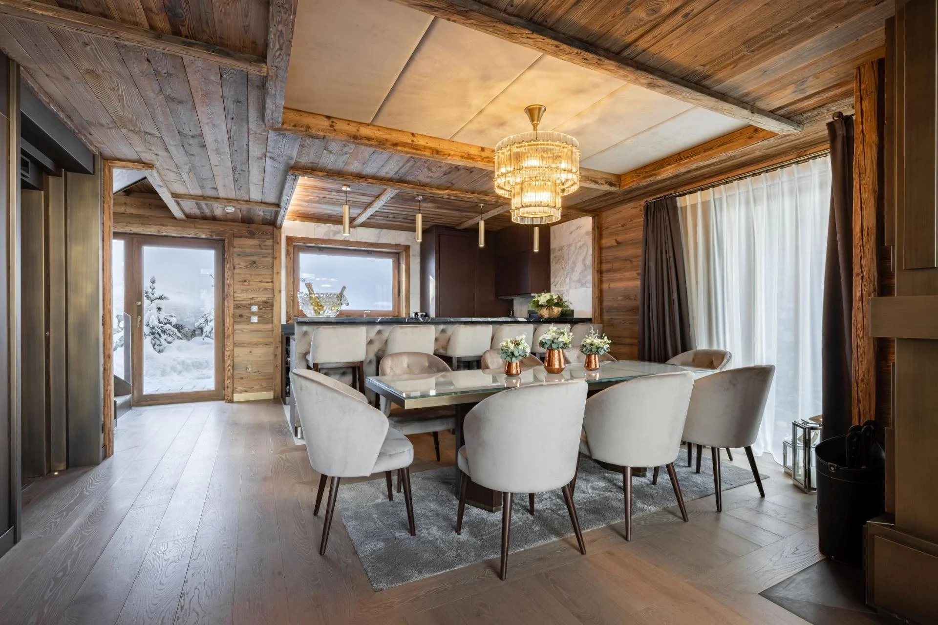 Interior of a rustic dining room featuring a large glass table surrounded by cream-colored chairs, wooden walls and ceiling, a chandelier, and a view of a snowy outdoor landscape through a glass door.