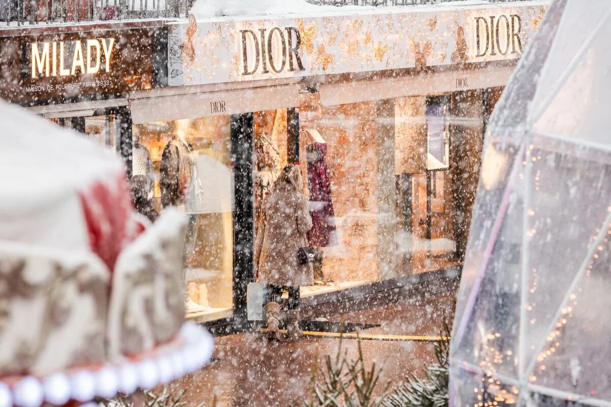 Courchevel 1850 storefront with a Dior sign, two mannequins displaying clothing, and a woman shopping in front of the window on a snowy day.