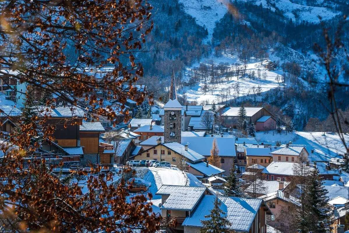 A snowy St Martin de Belleville village with houses, a church steeple, and snow-covered trees and hills in the background.