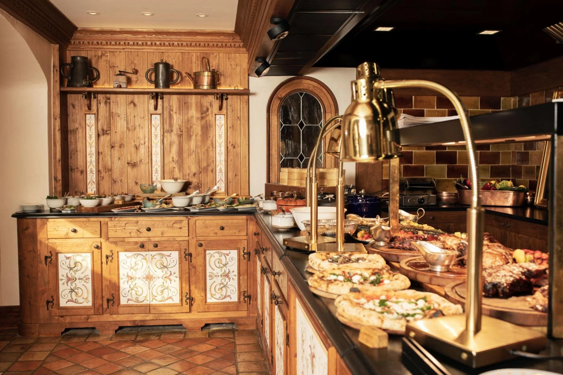 Interior of a rustic kitchen with wooden cabinets, a countertop filled with food and dishes, and a decorative backsplash. Gold-colored sink fixtures and a stained glass window are also visible.