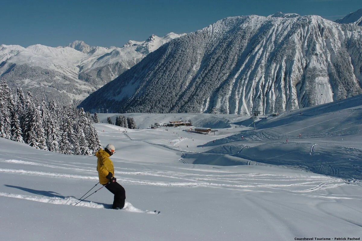 A skier in a yellow jacket and black pants skiing through fresh snow in Courchevel 1650, snowy landscape with snow-covered trees, a ski resort, and chairlifts in the distance under a clear blue sky.