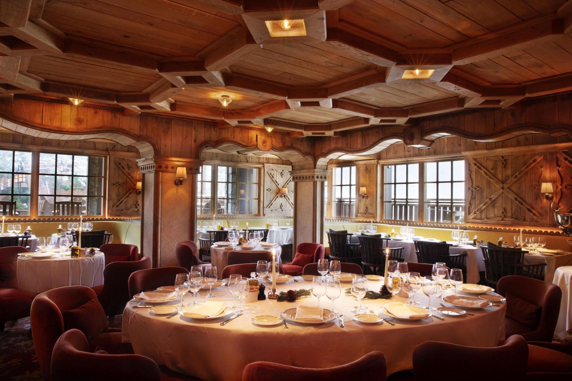Interior of a rustic wooden restaurant with various tables set for dining, including wine glasses and white tablecloths, large windows, and warm lighting.