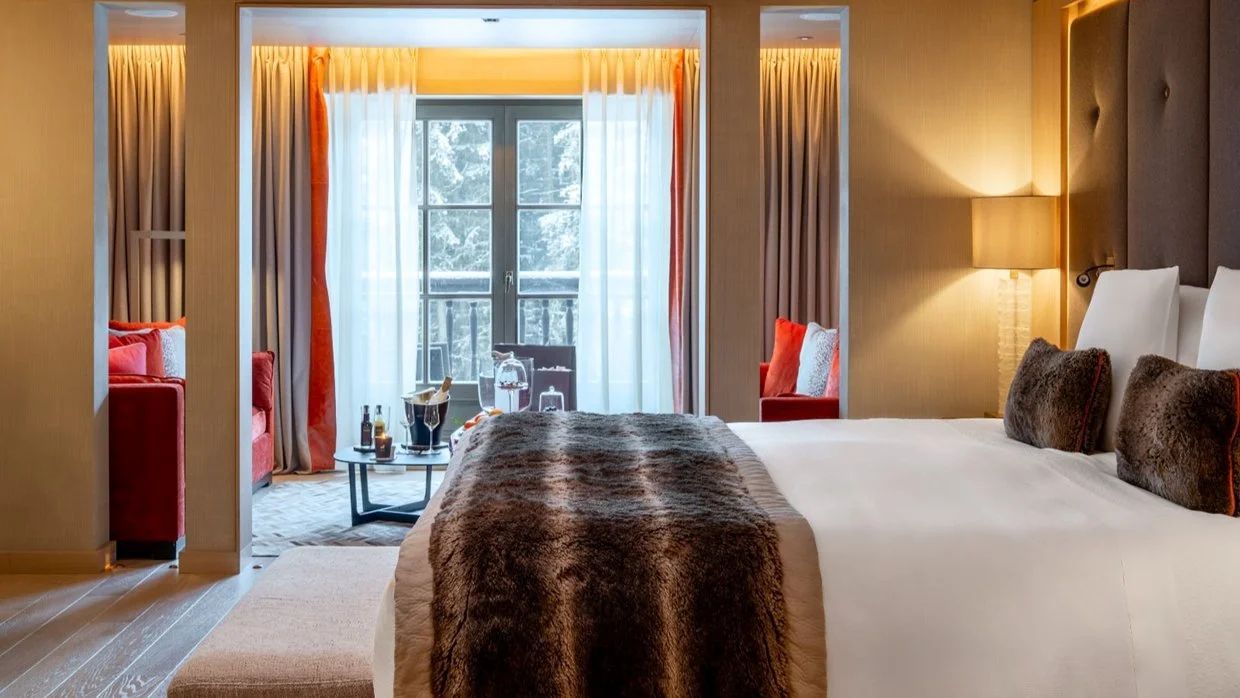 A cozy hotel room with a large bed and a fur throw, a bedside lamp, and seating area with red chairs and a small table in front of a balcony with snow-covered trees outside.