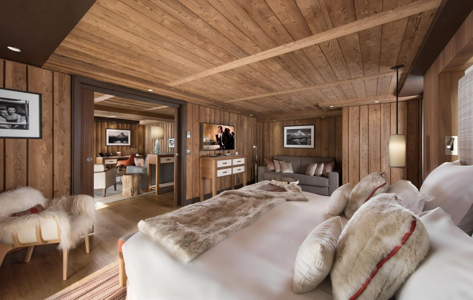 A cozy bedroom with wood-paneled walls and ceiling, featuring a bed with furry pillows and blankets, a small gray sofa, and a flatscreen TV mounted on a wooden dresser, with framed black and white photography on the walls.