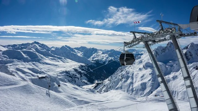 Snow-covered ski resort with ski lift cabins and mountain peaks under a blue sky with clouds.